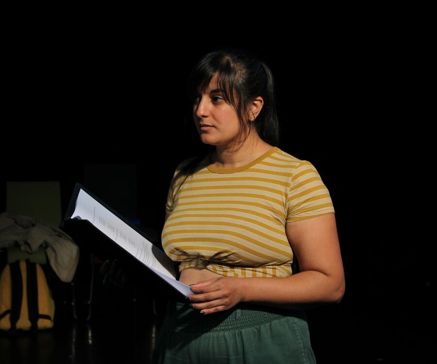 Stage photo of Michelle Kelly as a secretary, perched on a box and holding a clipboard