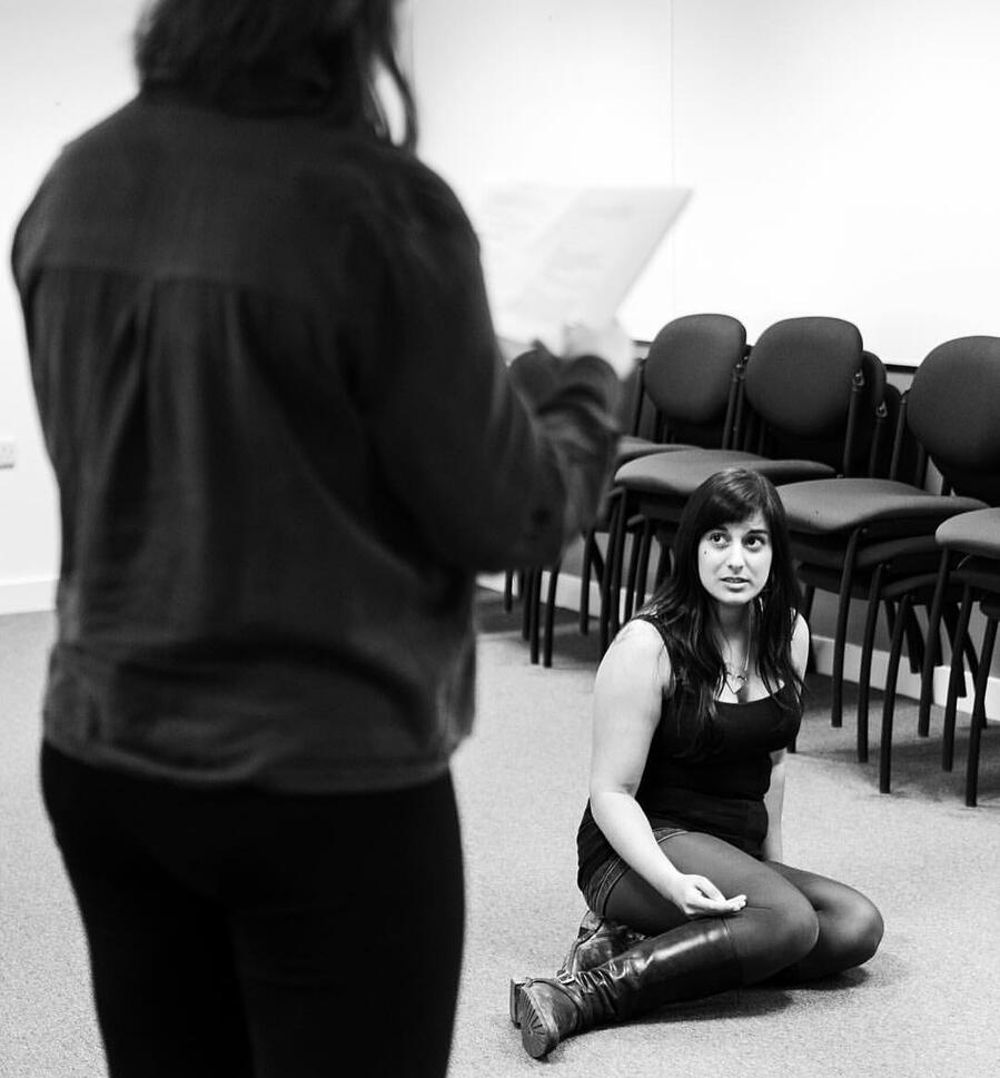 Rehearsal photo of Michelle Kelly sitting on the ground with a a cast member in foreground holding a script