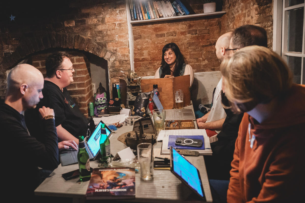 Chris Kendall Photography Photo of Michelle at one end of a table, with 5 players sat around smiling and listening to her. The table is covered in miniatures, dice, notebooks, drinks and snacks